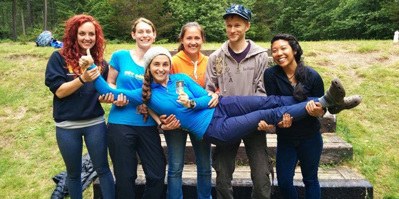 EarthCorps alum, Lindsey Falkenburg, being held by a group of smiling Corps Members. 