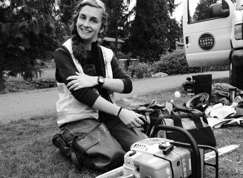 EarthCorps alum, Lindsey Falkenburg, sitting in the grass, smiling next to a chainsaw. 