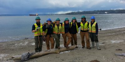 EarthCorps alum, Tom Lawler, and Corps Members smiling, wearing hard hats and safety vests on the beach.