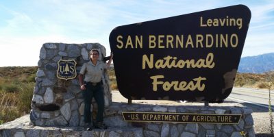 EarthCorps alum, Kayanna Warren, standing next to a sign that reads "Leaving San Bernardino National Forest. U.S. Department of Agriculture"
