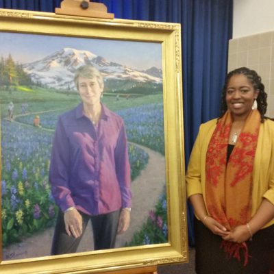 EarthCorps Alumna, Revina Moore, smiling and standing next to a painting of Sally Jewell.