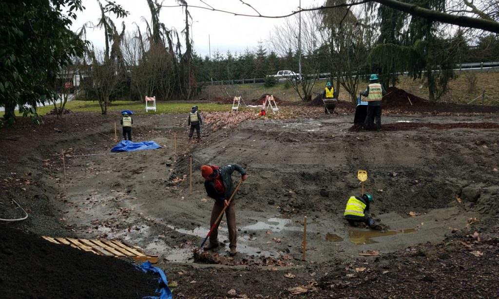 Corps Members shoveling and working in a large muddy, shallow hole.