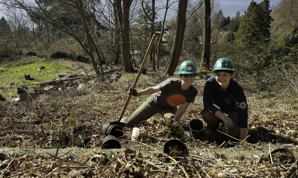 Two smiling Corps Members wearing hardhats and kneeling down next to plants, in an area surrounded by trees. One Corps member is holding a shovel.