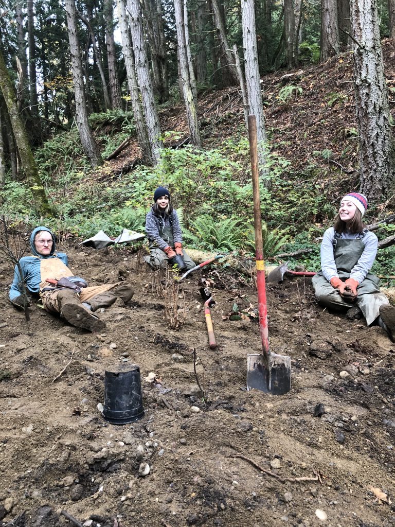 International Corps Members and EarthCorps staff sit in the dirt next to a shovel and plant pots.