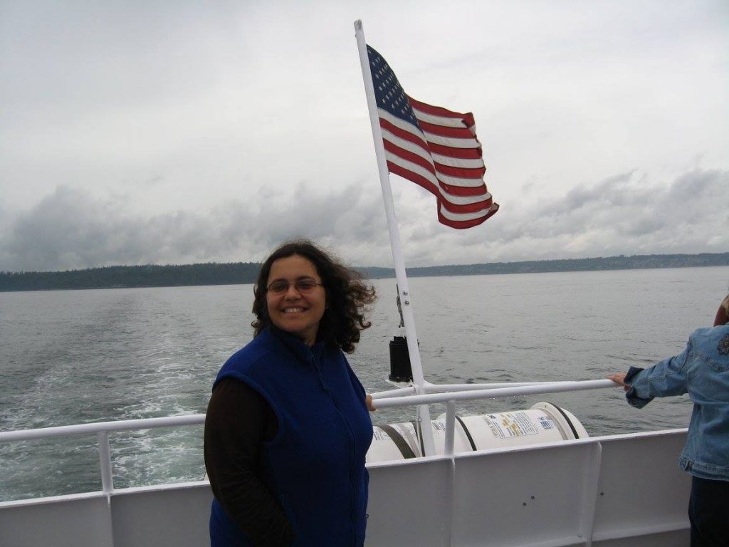 Mila standing at the back of a ferry boat, smiling at the camera as the wind blows an American flag behind her