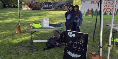 EarthCorps volunteer specialist, Nebras Maslamani, and a friend, standing in front of an EarthCorps sign in a grassy field at an Arabic Volunteer Event.