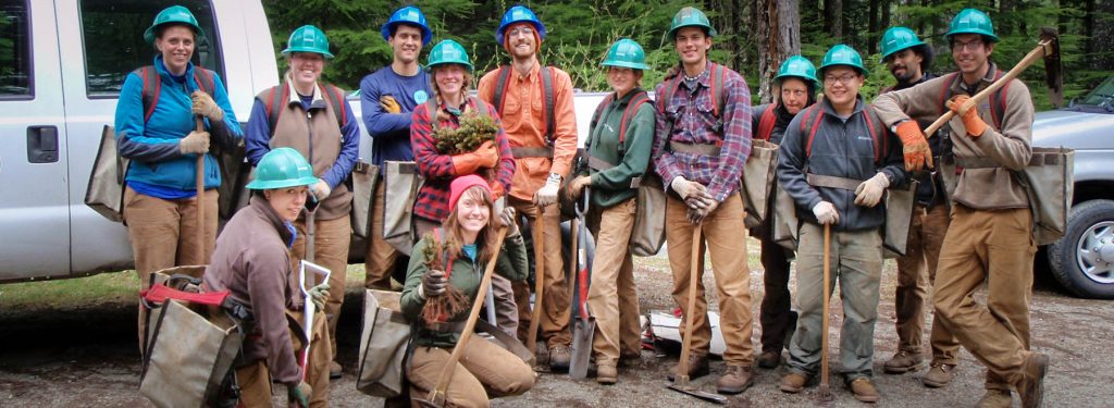 Group of smiling Corps Members, with backpacks, wearing hard hats and holding tools. 