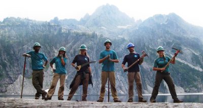 Group of Corps Members with hard hats on, holding tools and standing in front of mountains.