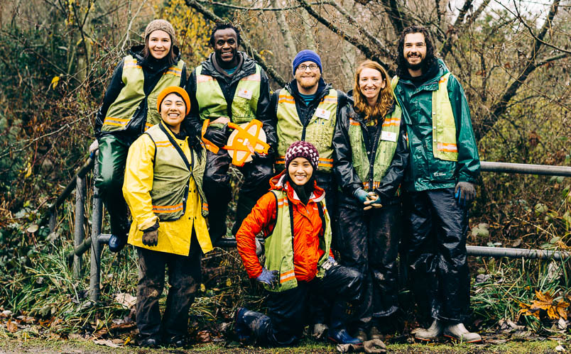 Group of smiling Corps Members wearing safety vests, standing in front of greenery.