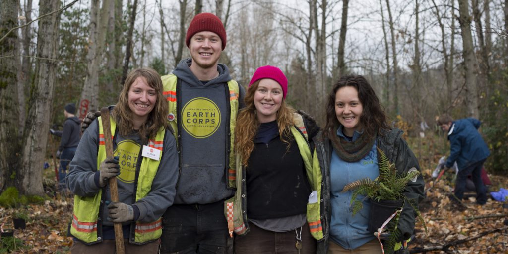 EarthCorps volunteer specialists smiling and holding plants and tools at a volunteer site.