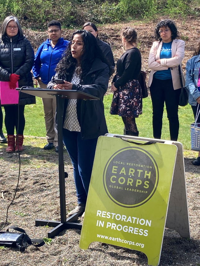 EarthCorps Executive Director, Evlyn Andrade, speaking in front in front of a podium and a group of people outdoors.