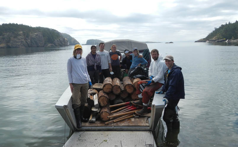 Smiling Corps Members on a boat with a stack of logs and several tools. 