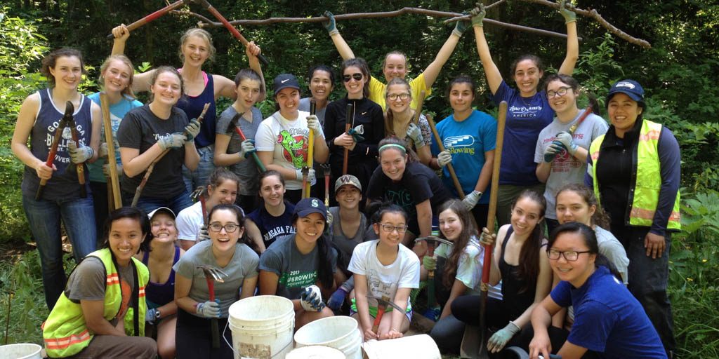 EarthCorps alum, Brittany Le with a large group of people all smiling and holding up tools. 