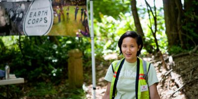 EarthCorps alum, Brittany Le smiling and wearing a safety vest, standing next to an EarthCorps sign outdoors.
