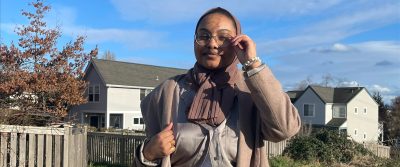 Iqra standing on the road in front of a light grey house. There are blue skies and she's smiling at the camera while touching round glasses.