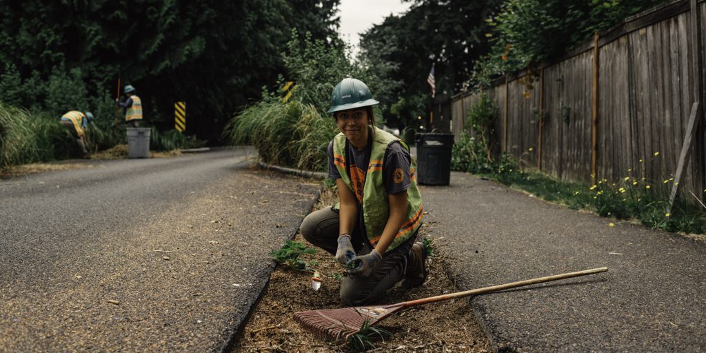 Smiling Corps Member wearing a hard hat and safety vest, kneeling down next to a rake at a field site.