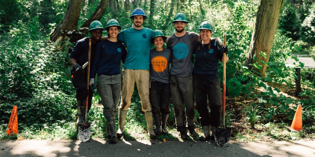 A group of smiling Corps Members wearing hard hats and holding shovels, standing in front of a forested area. 