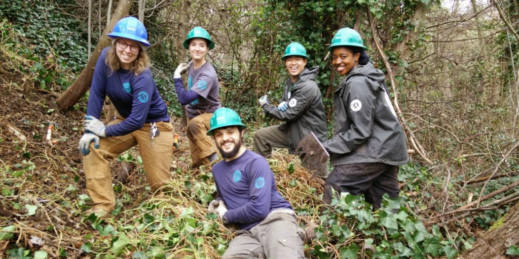 Corps Members wearing hardhats smiling next to a pile of ivy that had been removed.