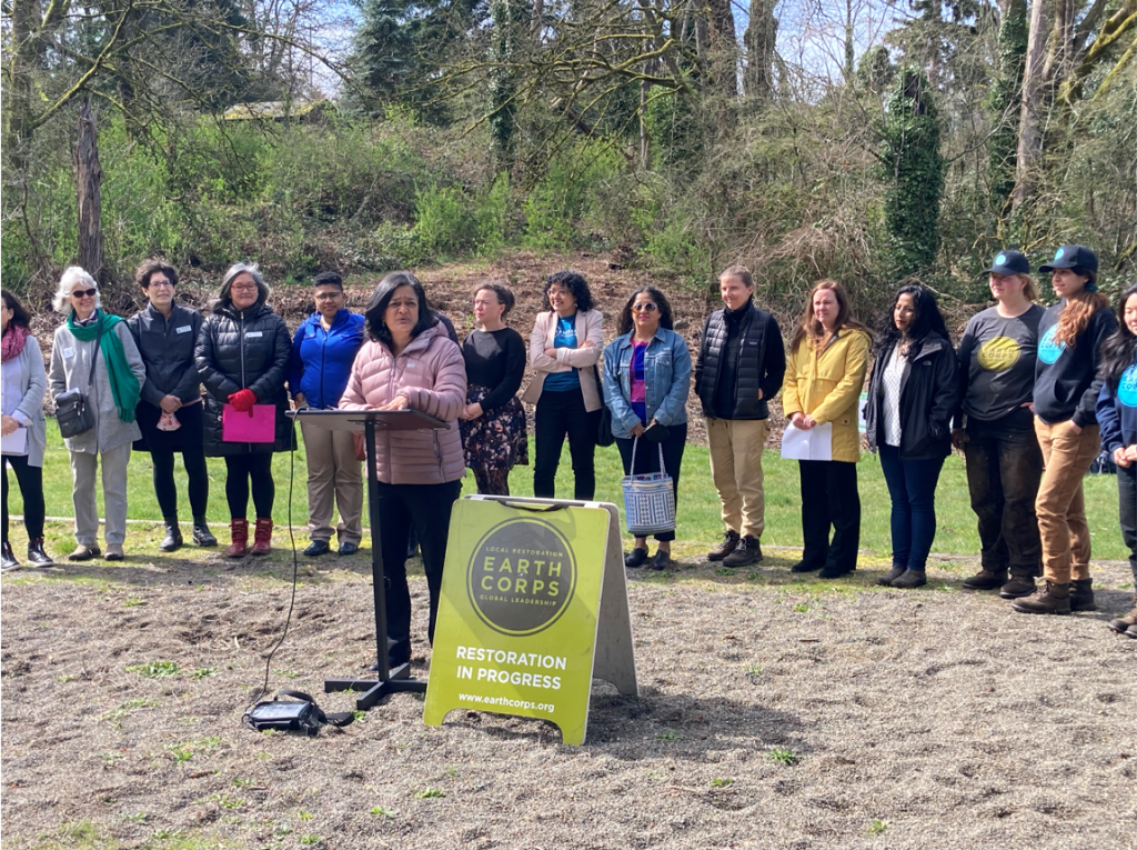 Congresswoman, Pramila Japyapal, speaking in front in front of a podium and a group of people outdoors.