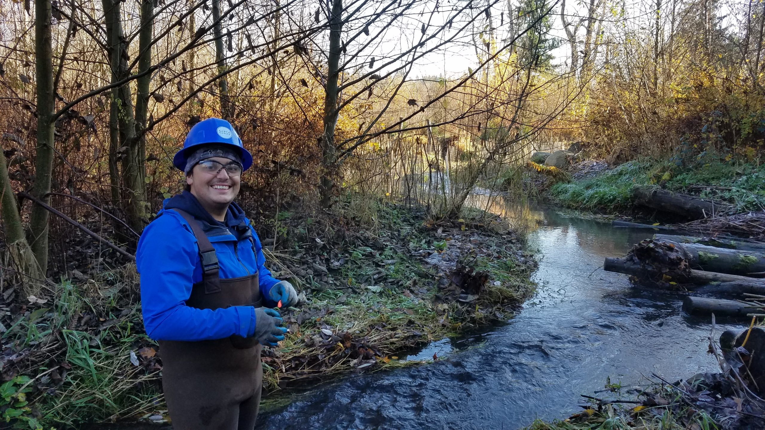 Smiling Corps Member wearing a hard hat and waders, standing in front of a small flowing body of water in a forested area.