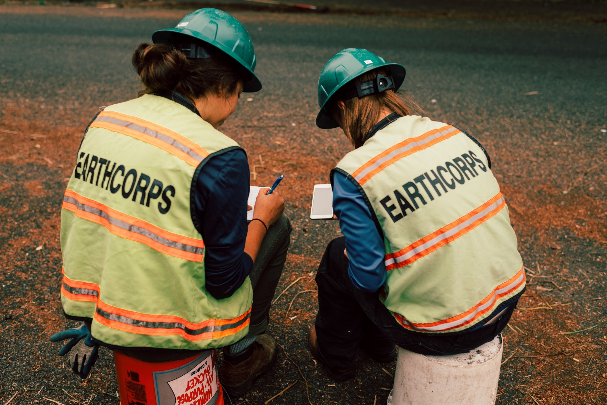 Two Corps Members wearing hard hats and safety vests, sitting down on buckets and writing on paper.