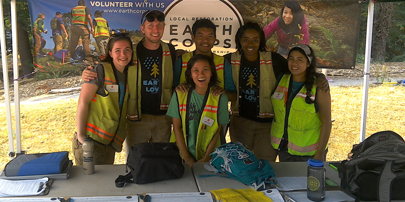 Alumna, Revina Moore smiling with a group of Corps Members wearing safety vests, in front of an EarthCorps sign. 