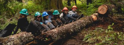 Six EarthCorps crew member sitting on the ground and pushing a large log with their legs while laughing.