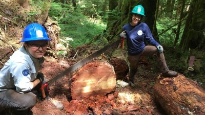 Two smiling Corps Members holding a long saw on a large log that has been cut.