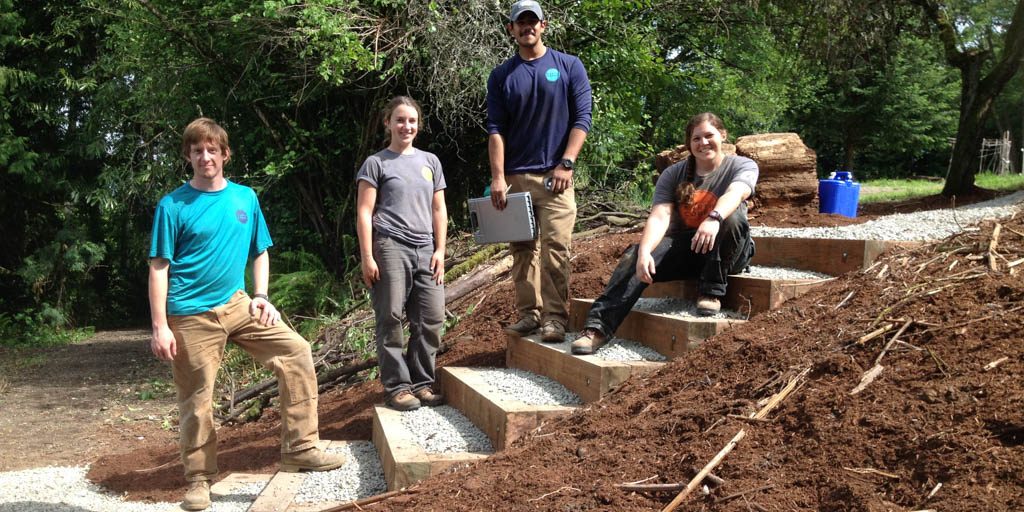 EarthCorps alum, Nicole Marcotte and three Corps Members smiling and standing on trail stairs.