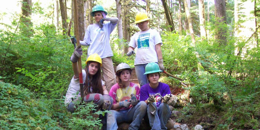 Volunteer Specialist, Natalie Izzo and International Corps Member, Hala, with a group of students wearing hard hats and holding tools outdoors in a forested area. 