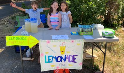 Three children smiling while standing at their lemonade stand