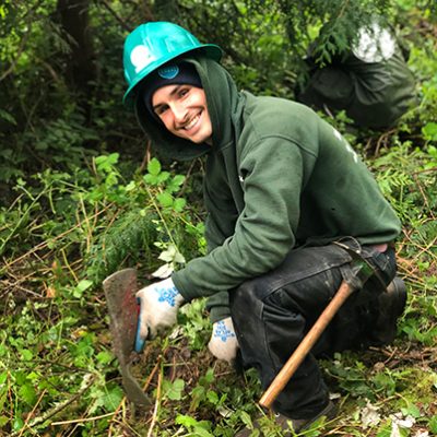 Smiling Corps member bending down in patch of green space with EarthCorps hard hat on