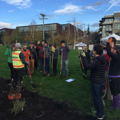 Volunteers outdoors holding shovels and listening to Morgan from EarthCorps speak. 